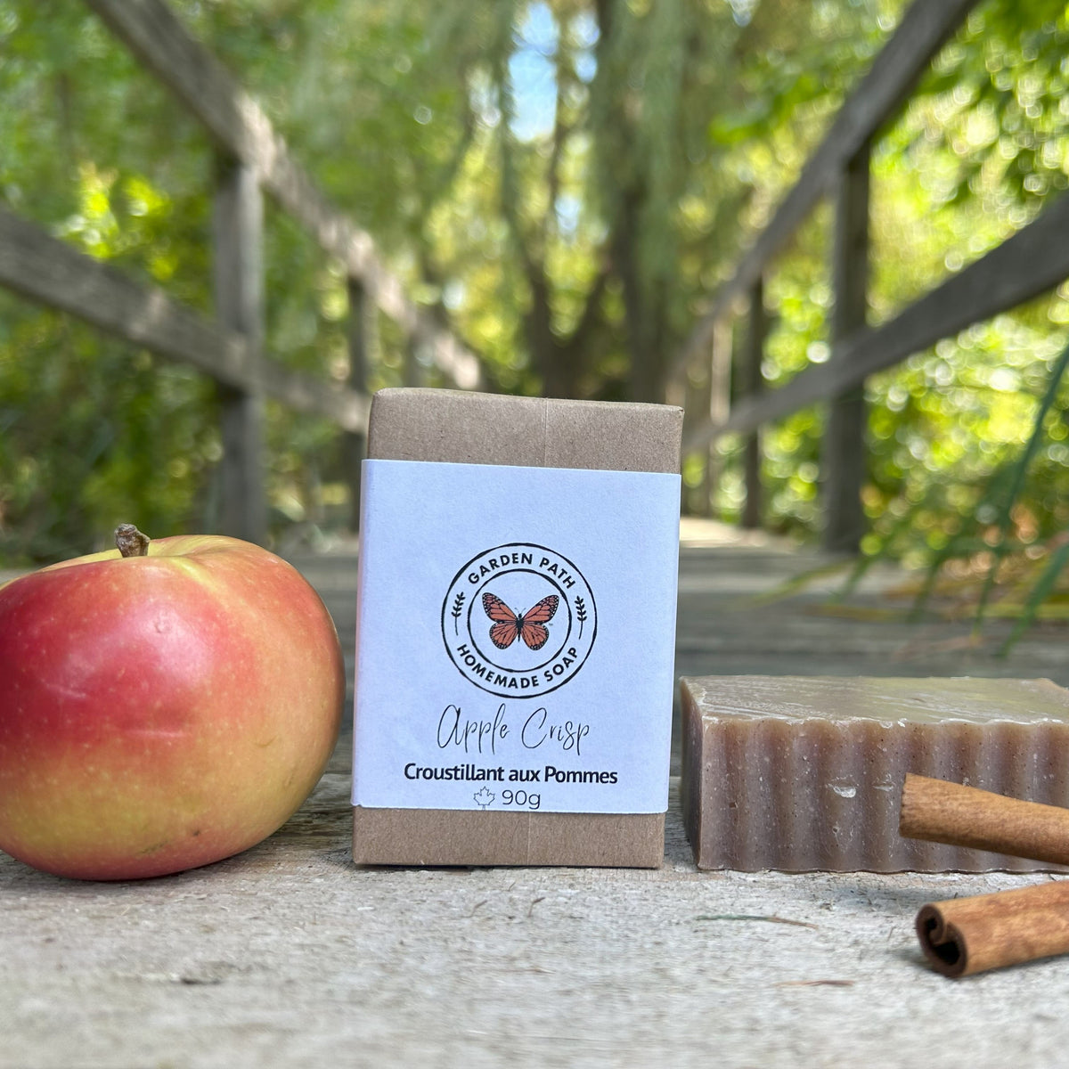 Bar of soap with packaging labeled 'Apple Crisp' next to an apple and cinnamon sticks outdoors by Garden Path Homemade Soap. Exfoliating with Spicy Fragrance Oils