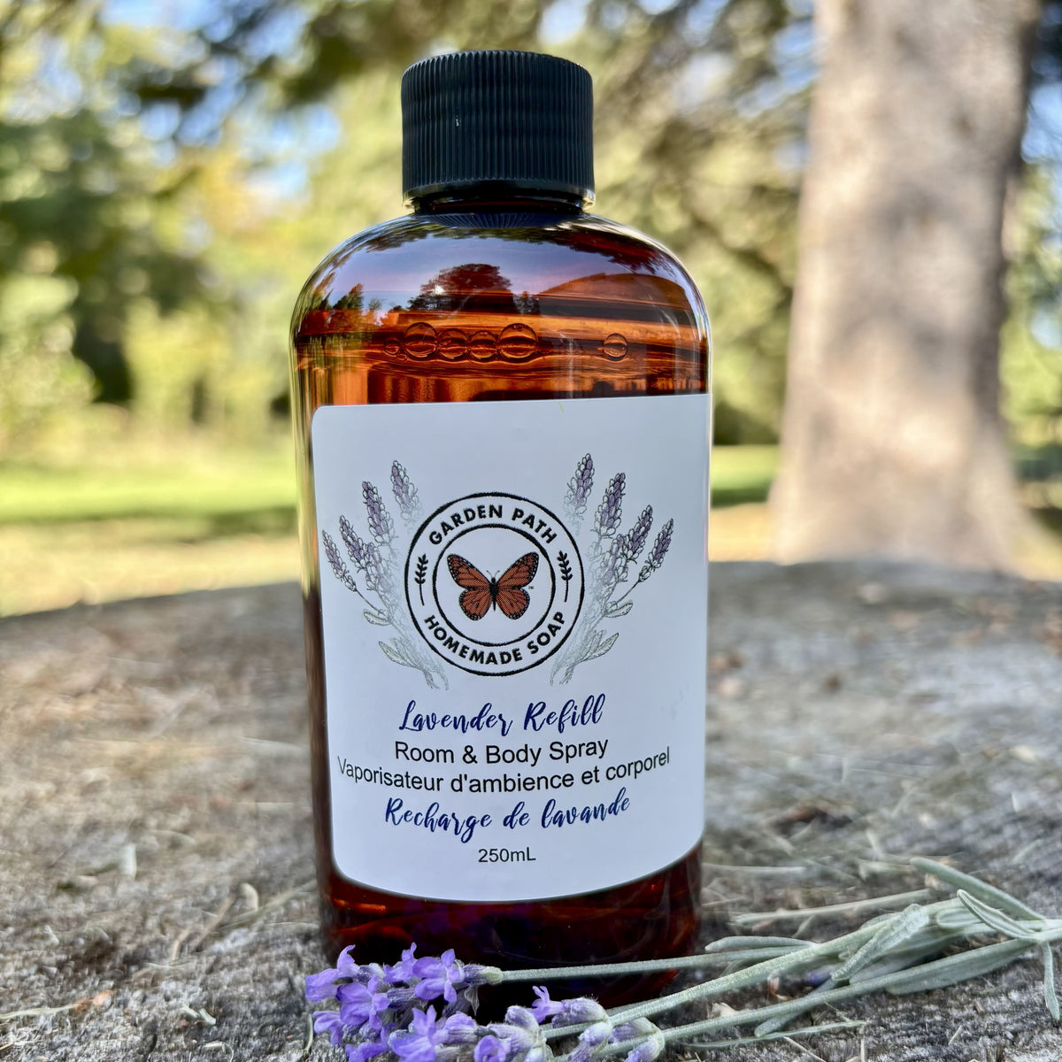 Bottle of lavender refill room and body spray sitting on a wooden surface with lavender flowers by Garden path Homemade Soap. 100% Natural Essential Oil