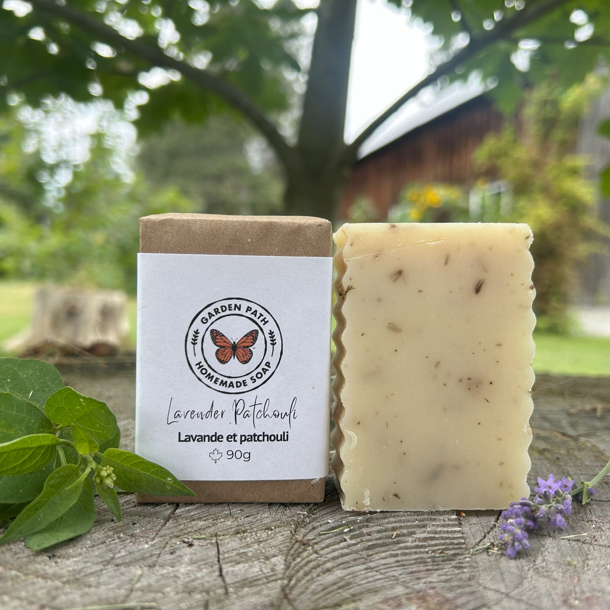 Lavender Patchouli Bar of soap with a label on a wooden surface outdoors. One bar wrapped the other unwrapped. Made with 100% Natural Essential Oils and Lavender Buds by Garden Path Homemade Soap