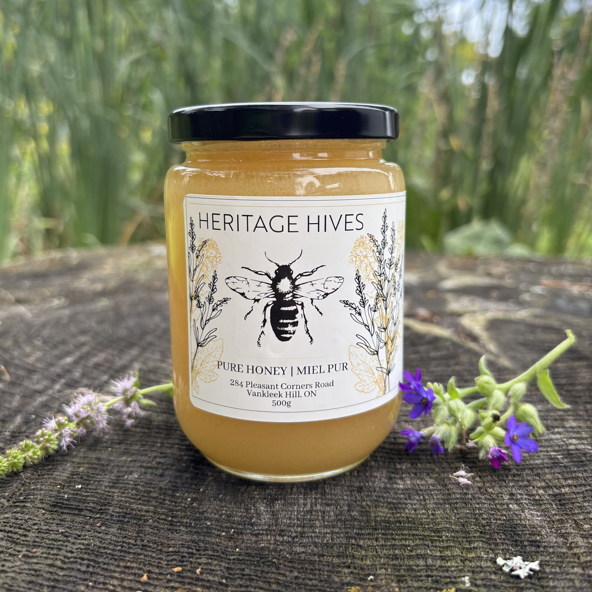 A jar of Heritage Hives Pure Honey with a black lid and white label, set on a dark wooden surface outdoors with green foliage in the background and small purple flowers in the foreground.