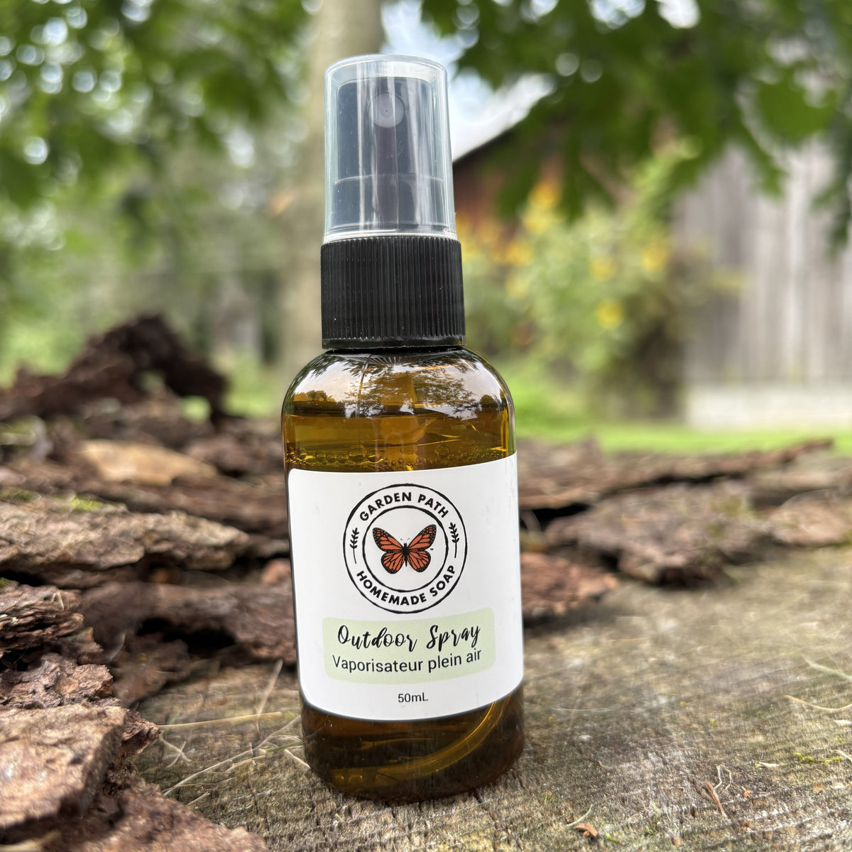 A 50mL amber glass spray bottle of Garden Path Outdoor Spray with a black nozzle and clear cap, resting on a rustic tree trunk with a blurred outdoor garden background. 100% Natural Essential Oil Blend - Garden Path Homemade Soap