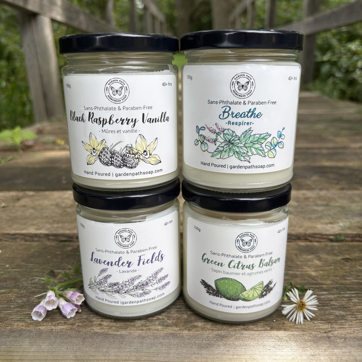 Four large soy candles arranged in a 2x2 grid on a rustic wooden table. The background shows a blurred outdoor garden setting. Garden Path Homemade Soap
