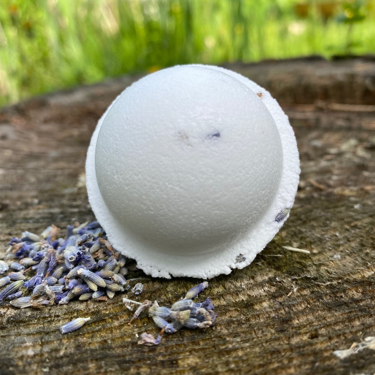 A white lavender bath bomb with lavender buds visible on the surface, placed on a wooden surface with a scattering of lavender buds around it by Garden Path Homemade Soap. 100% Natural Essential Oil & Ingredients