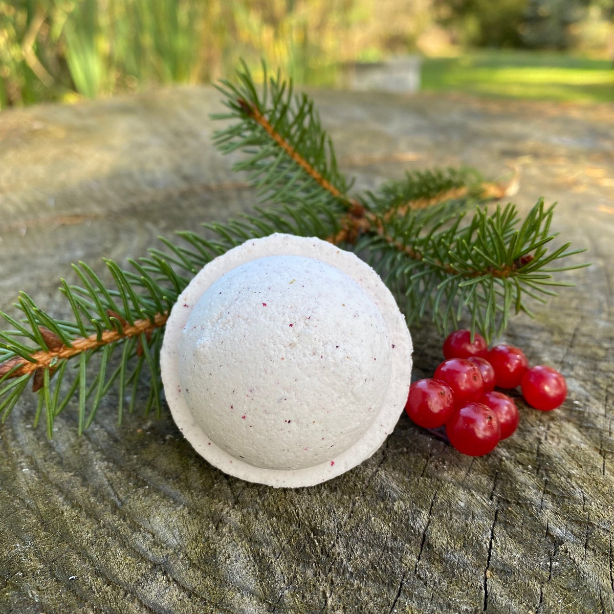 Frosted Winterberry Bath Bomb with a frosted white exterior and red speckles, placed on a wooden surface with green pine needles and red berries to the side.