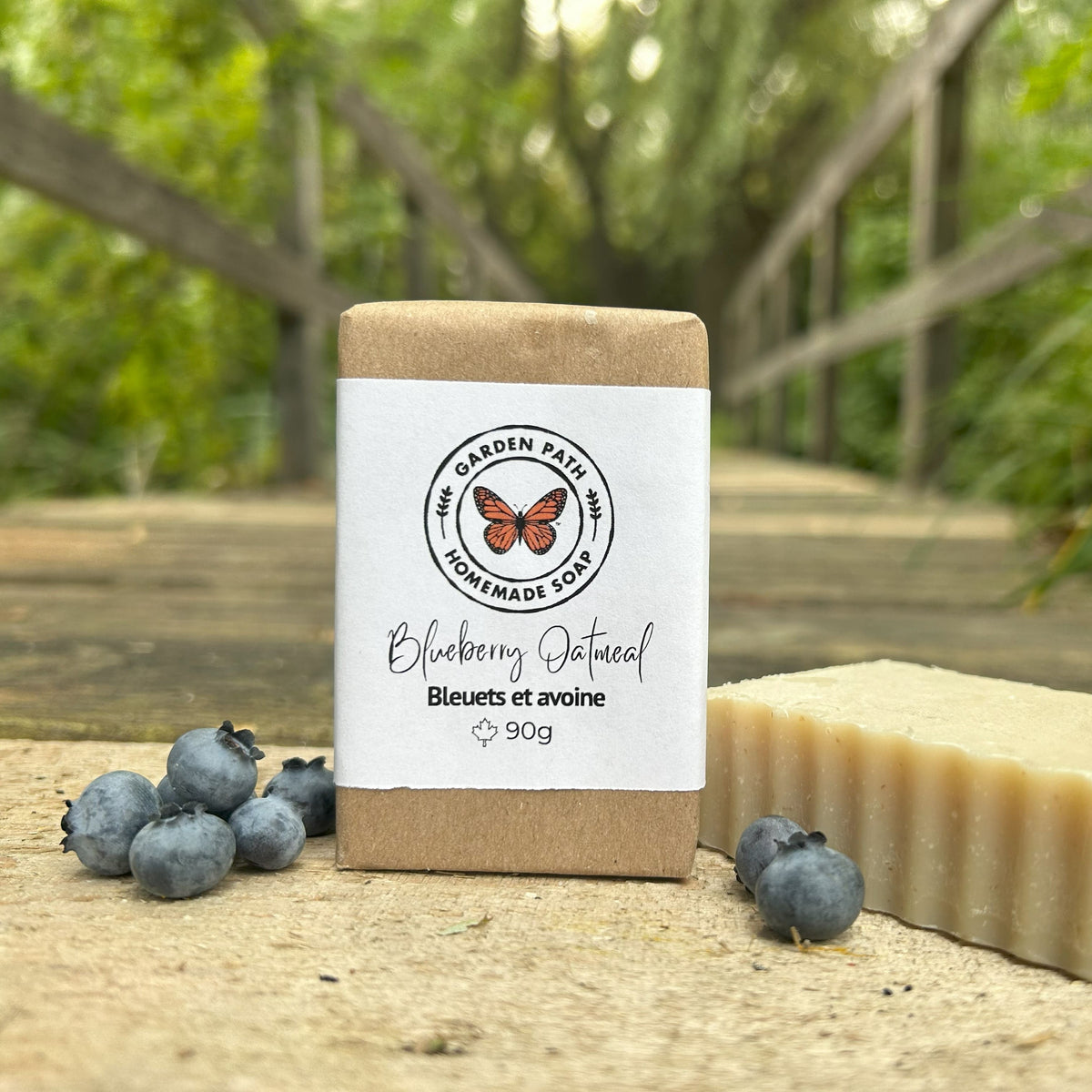 Blueberry Oatmeal Bar Soap both wrapped and unwrapped with blue berries scattered around on a wooden surface with natural green background. Naturally Exfoliating Soap Scented with Fragrance oil - Garden Path Homemade Soap