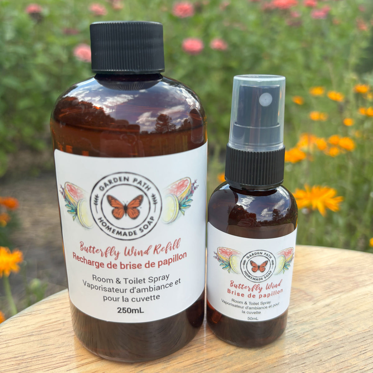 Butterfly Wind 50ml spray and 250ml refill bottle - Room & Toilet Spray by Garden Path Homemade Soap on a wooden surface with flowers in the background. 100% Natural Essential Oil Blend