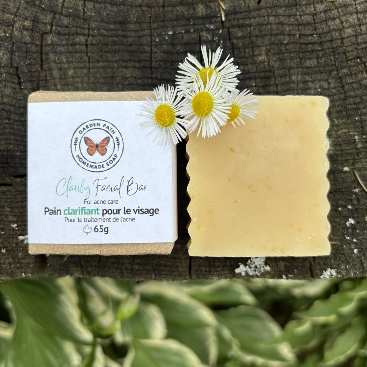 A handmade facial bar soap Clarify Facial Bar for acne care shown next to its paper packaging, placed on a rustic dark wood surface with white  flowers on top.  100% Natural Ingredients - Garden Path Homemade Soap