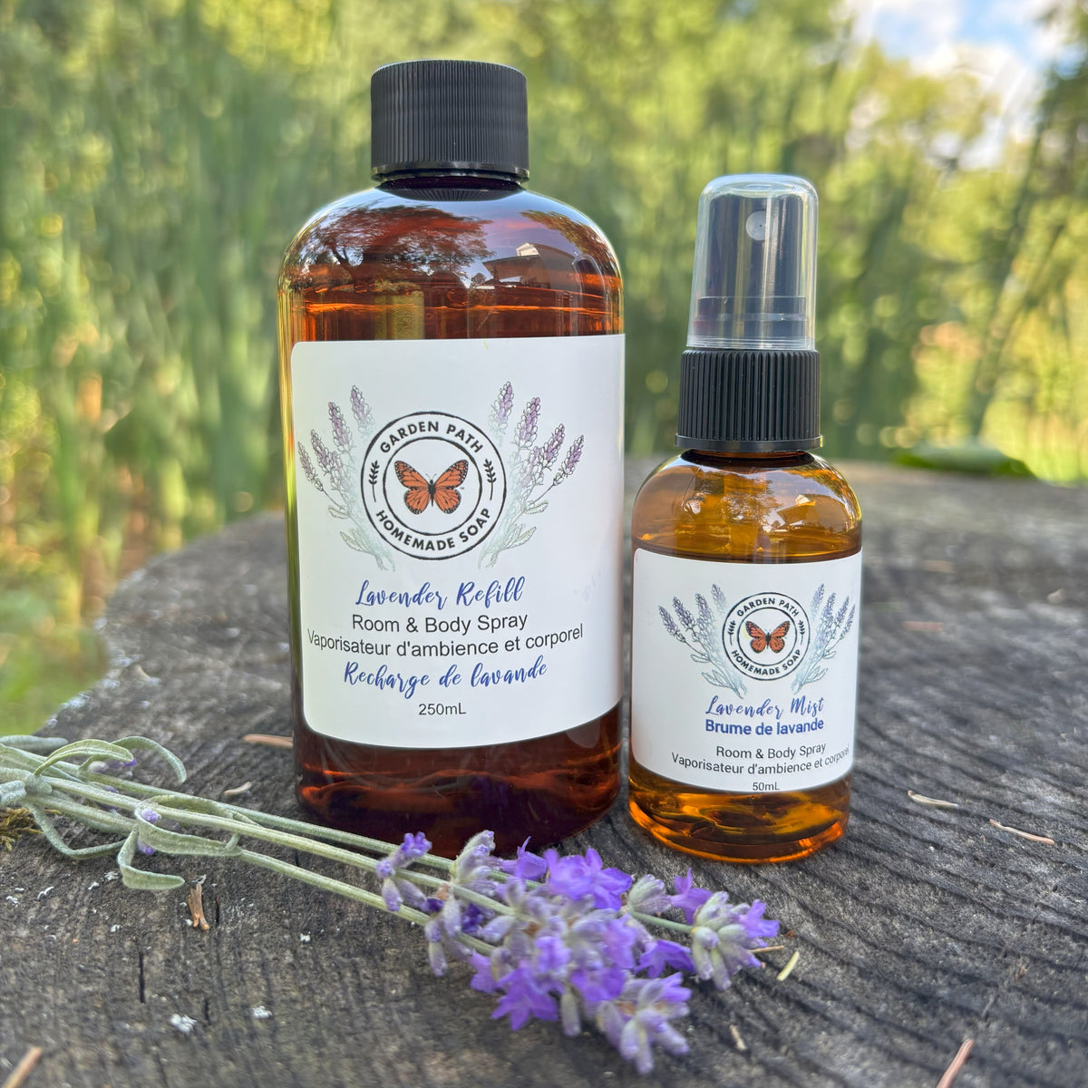Two bottles of lavender-scented room, one a spray bottle and one a refill bottle  on a wooden surface with lavender flower by Garden Path Homemade Soap.  100% Natural Essential Oil