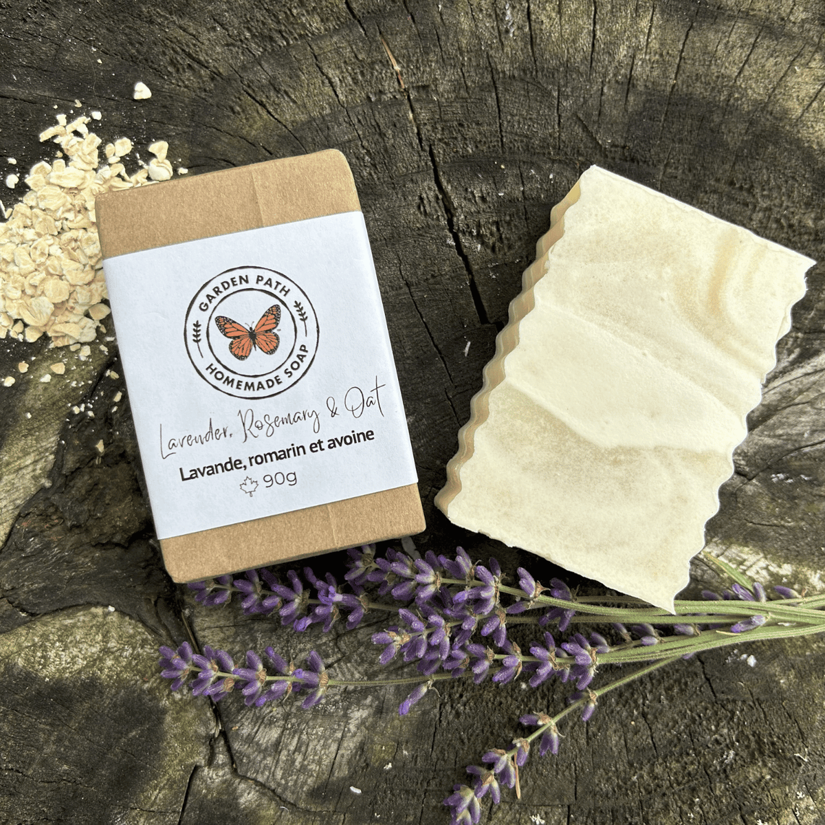 A 90g bar of handmade soap by Garden Path Homemade soap  labeled 'Lavender, Rosemary & Oat' sits next to its kraft paper packaging on a rustic wood surface, styled with dried lavender flowers and oats.