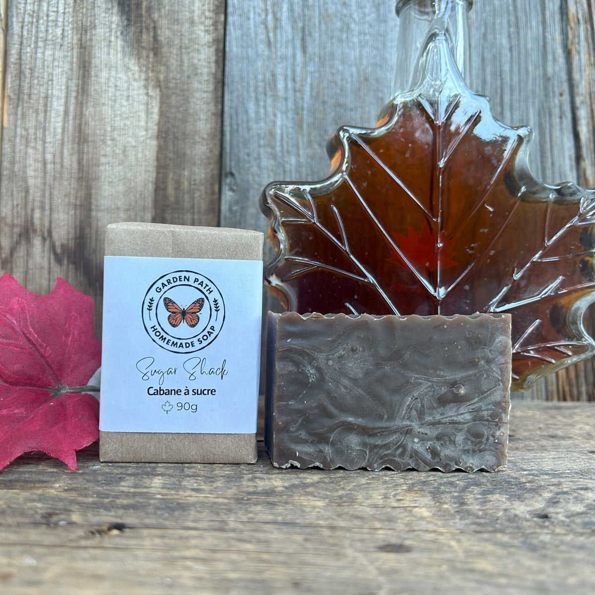 Garden Path Homemade Soap's Sugar Shack Bar of soap with packaging and a maple leaf glass container filled with maple syrup  on a wooden surface. Lightly Scented Maple Fragrance & Natural Maple Sugar Sand