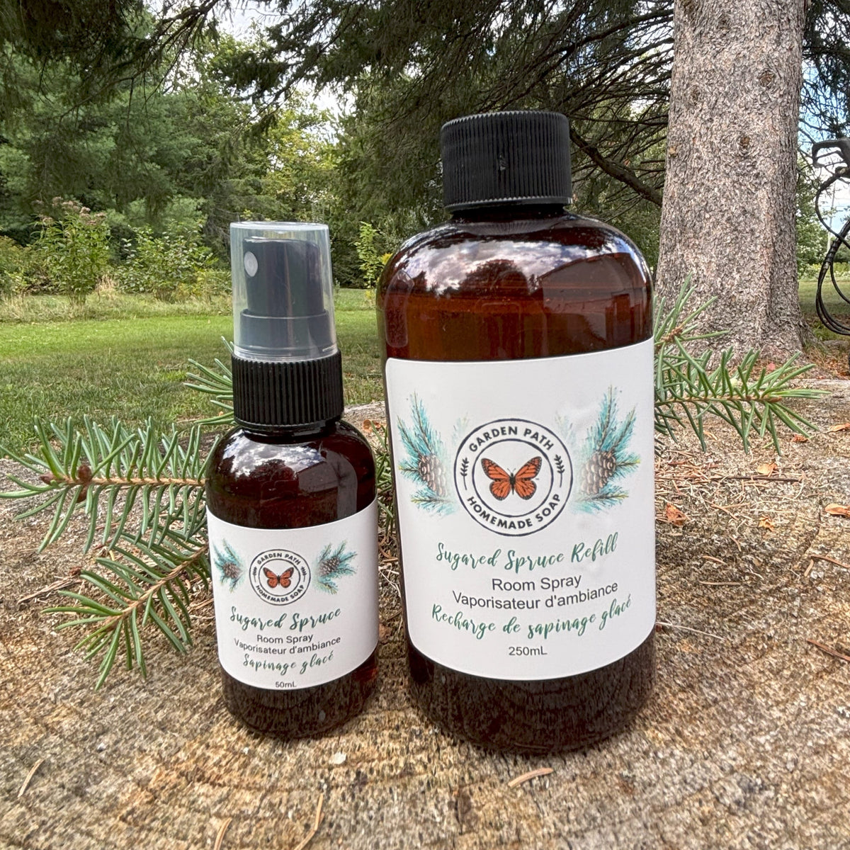 Sugared Spruce Fragrance Oil in spray bottle and refill bottle sitting on wooden surface with a spruce branch beside and green trees in the background.