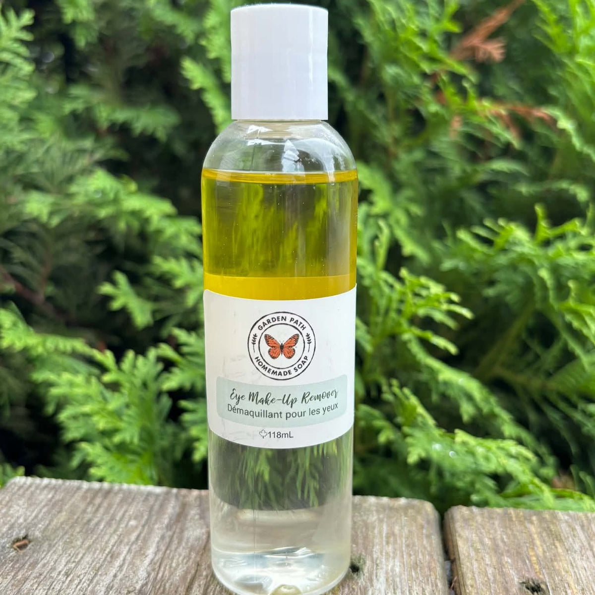 Eye make up remover bottle sitting on a wooden surface with cedar tress in the background by Garden Path Homemade Soap. 100% Natural Ingredients