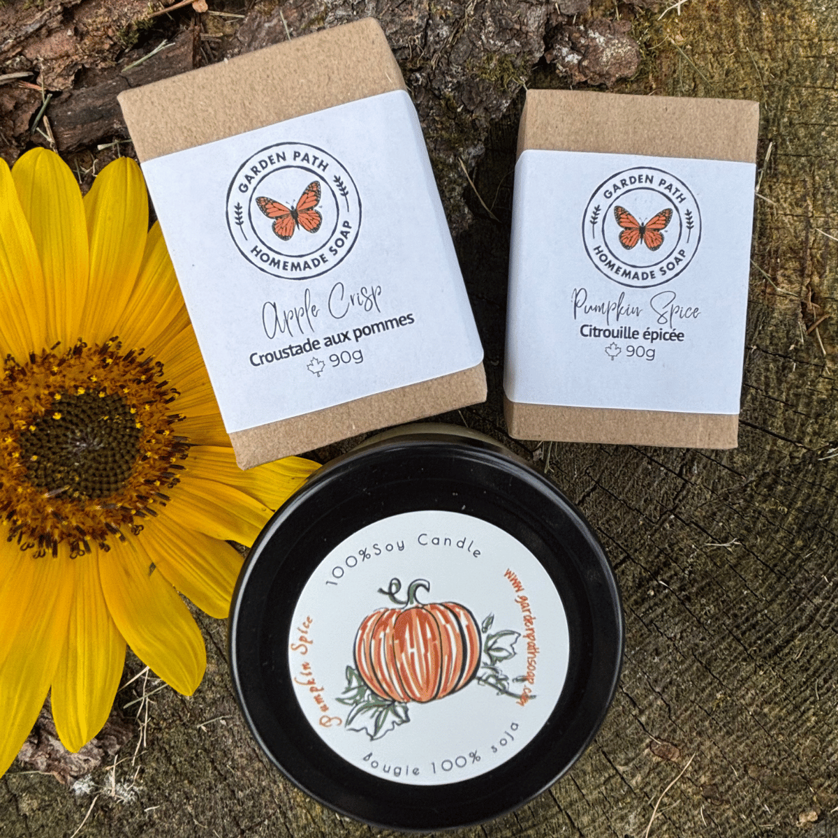 Apple Crisp and Pumpkin Spice soap bars and one small candle of pumpkin spice  from Garden Path Homemade Soap are arranged on a rustic wood surface with a yellow sunflower. 