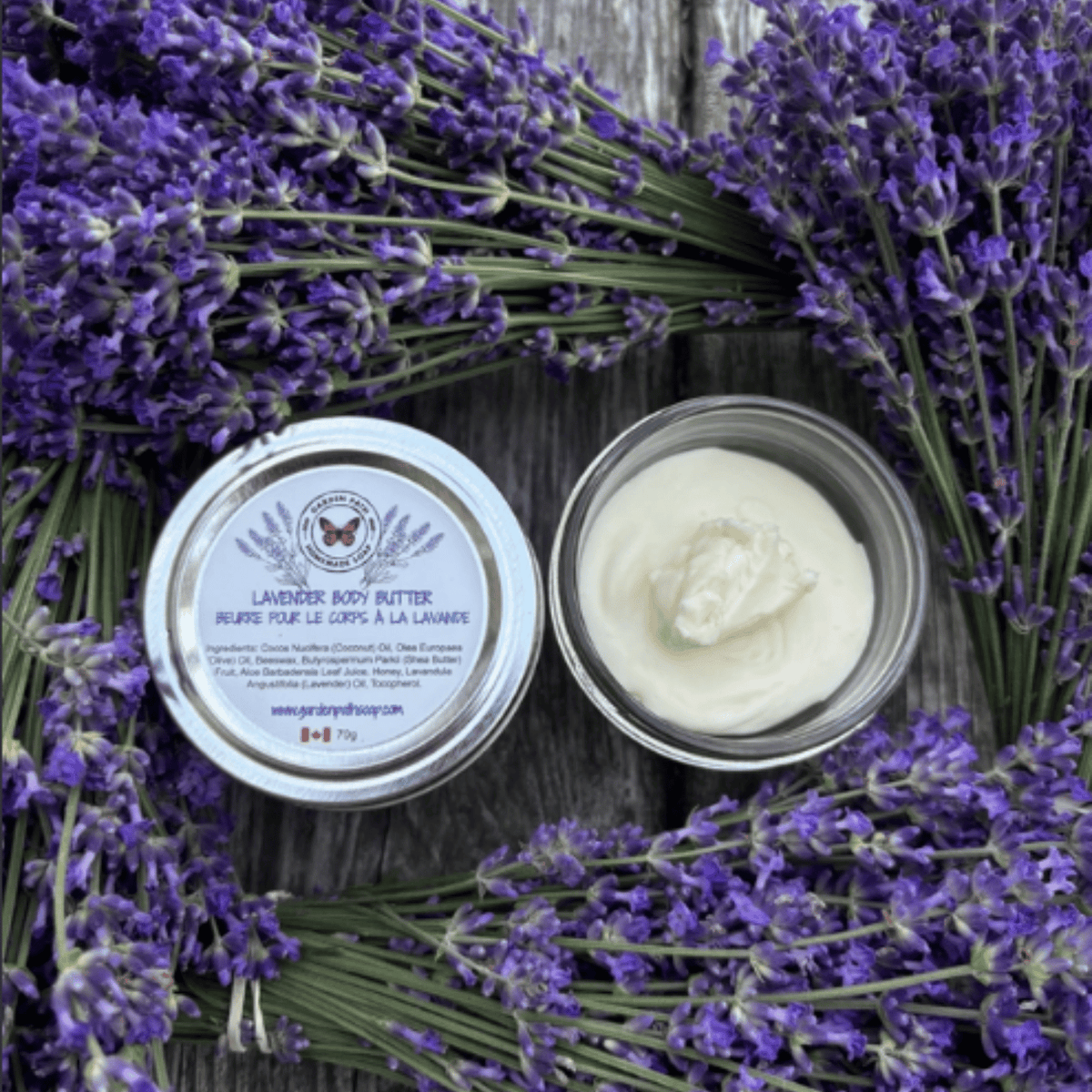 Lavender body butter in a clear round jar with a white lid labeled 'Lavender Body Butter' by  Garden Path Homemade soap, placed on a rustic wooden surface surrounded by fresh purple lavender sprigs.