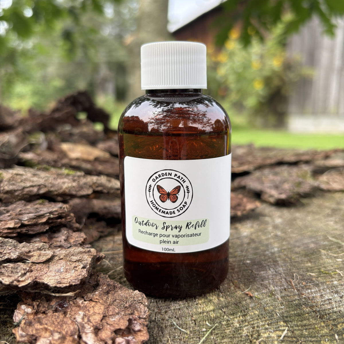 A 100ml amber glass bottle of  Outdoor Spray Refill with a white cap and white label, placed on a rustic log with a blurred outdoor garden background.  100% Natural Essential Oil Blend - Garden Path Homemade Soap