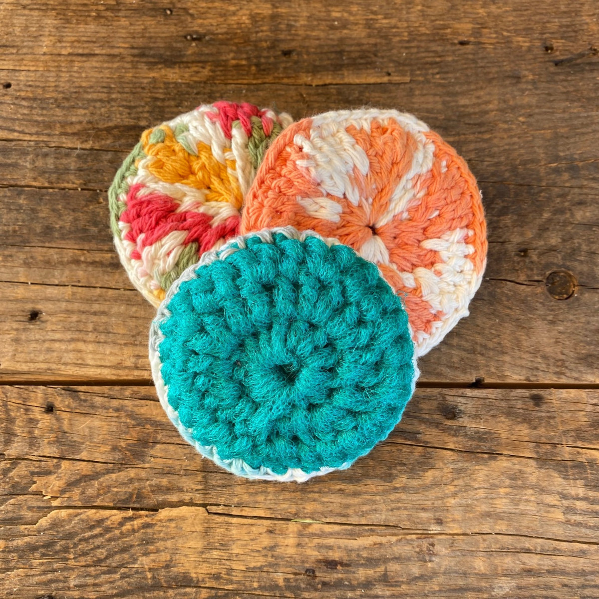 Three handcrafted, crocheted pot scrubbies in multicolor, orange, pink, and teal, displayed on a wooden surface.